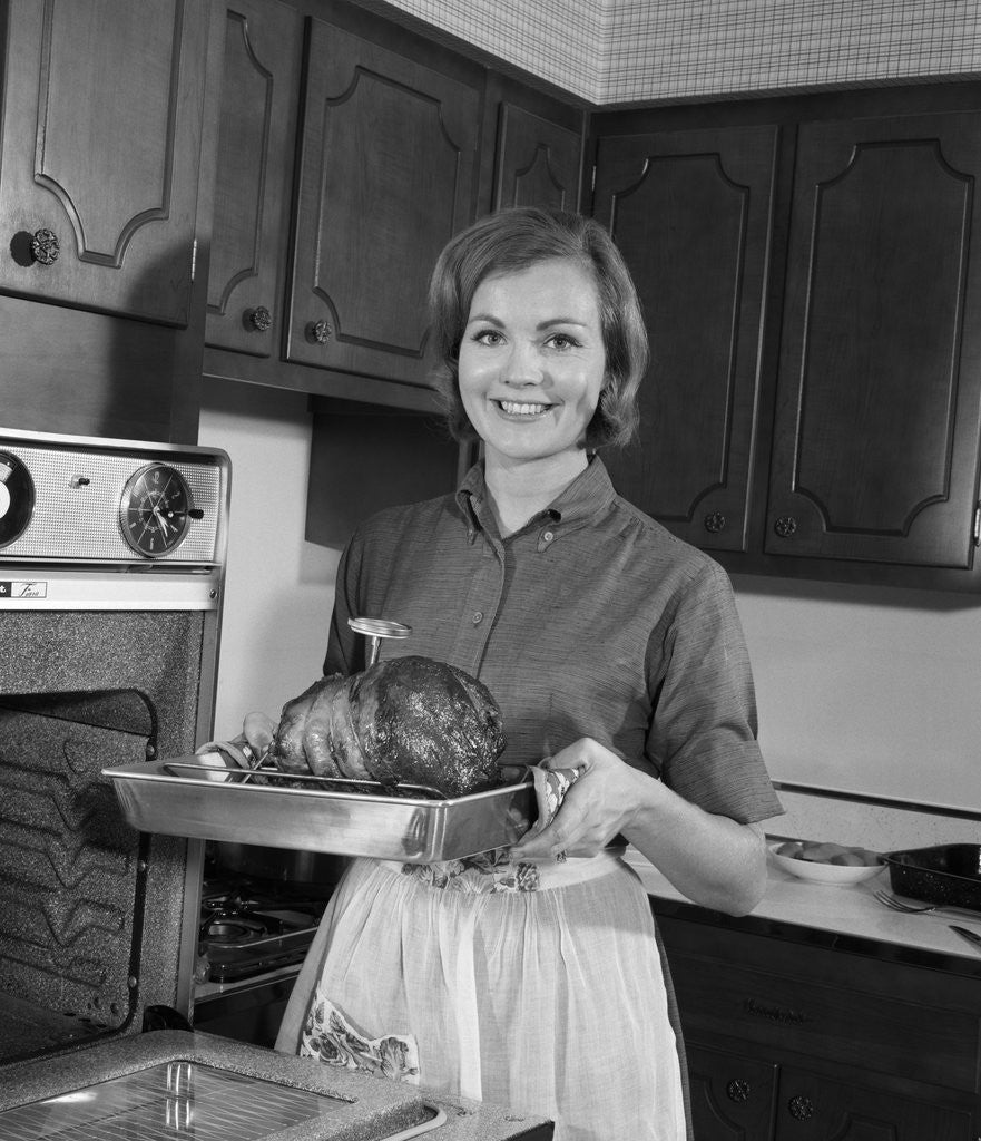 Detail of 1960s smiling woman in kitchen taking roast out of oven looking at camera by Anonymous