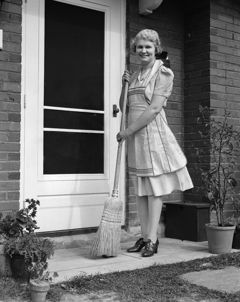 Detail of 1940s smiling woman sweeping porch front door step with a broom looking at camera by Anonymous