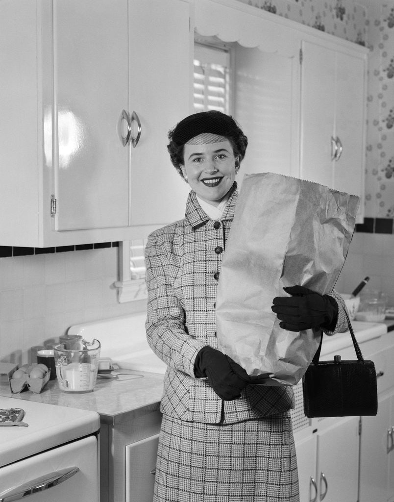 Detail of 1950s smiling woman in kitchen holding grocery bag handbag wearing hat gloves looking at camera by Anonymous