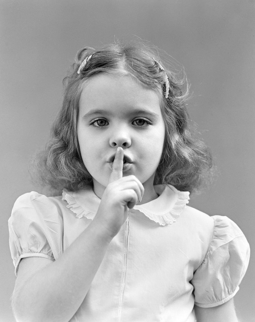 Detail of 1940s 1950s girl with finger to lips making quiet shush gesture looking at camera by Anonymous