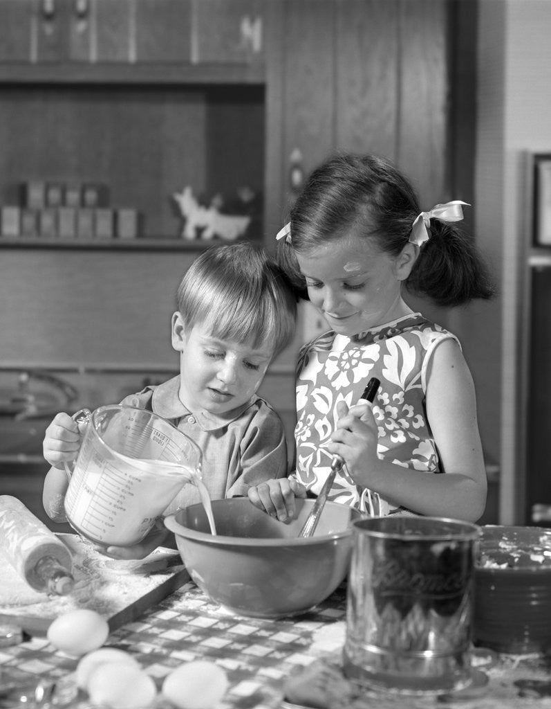 Detail of 1960s two children boy girl bowl mixing pouring milk in kitchen by Anonymous