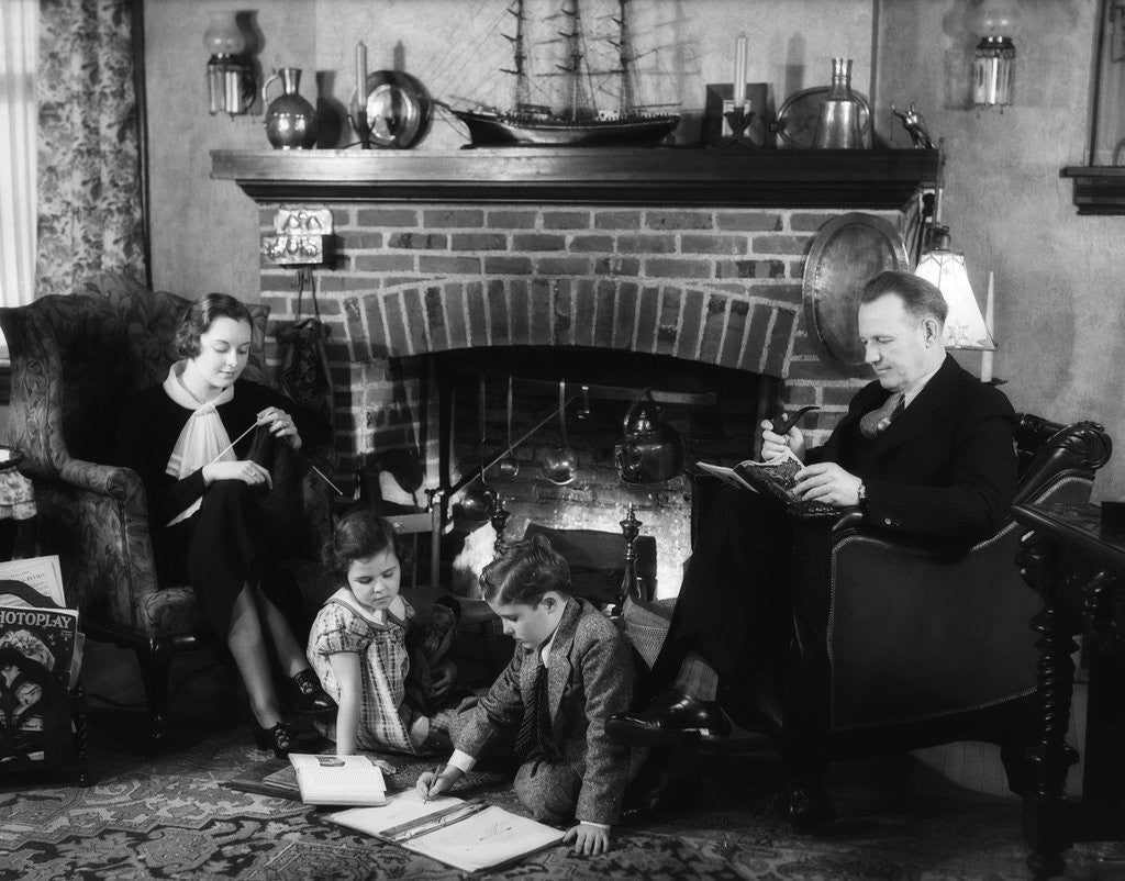 Detail of 1930s family of four sitting in front fireplace mother knitting father reading smoking pipe kids doing homework by Anonymous