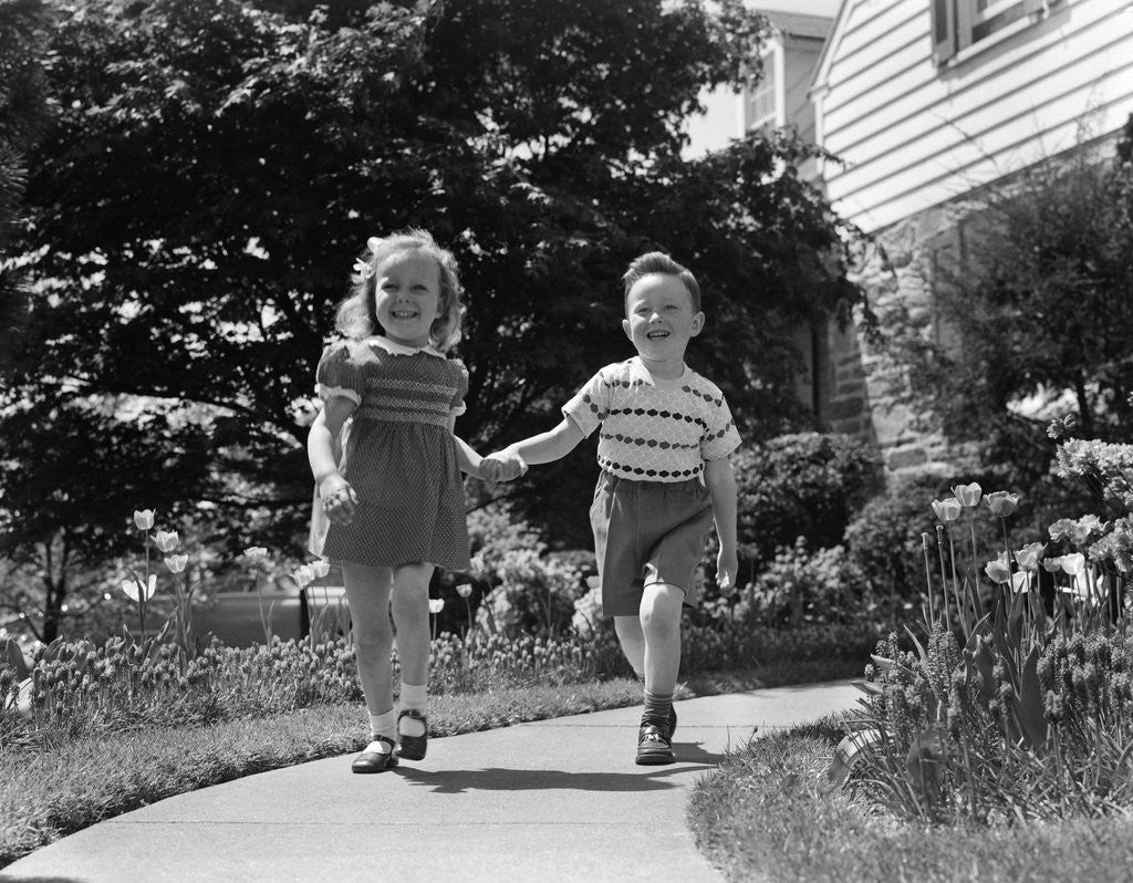 Detail of 1950s two children holding hands smiling walking along sidewalk by Anonymous