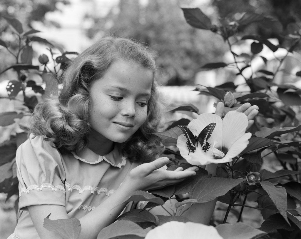 Detail of 1950s pretty little girl smiling at hibiscus flower & butterfly in garden by Anonymous