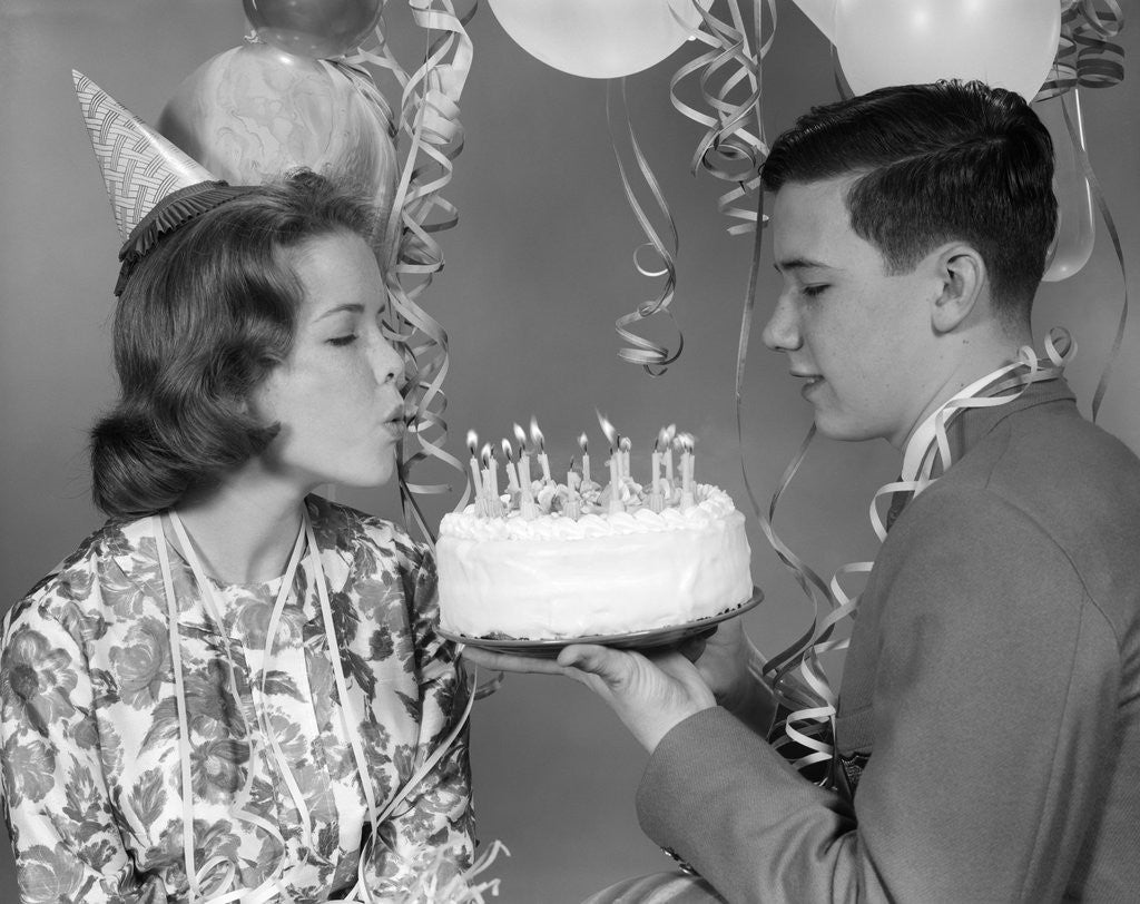 Detail of 1960s teenage girl blowing out candles on birthday cake held by boy by Anonymous