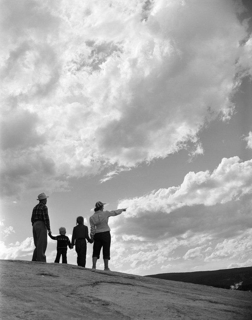 Detail of 1950s 1960s family of four silhouetted on top rocky hill pointing at clouds in sky by Anonymous