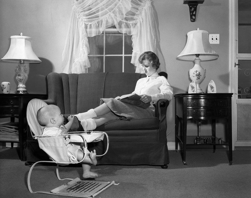 Detail of 1950s teen babysitter seated on sofa reading schoolbook while baby in bouncy sling chair grabs her propped up foot by Corbis