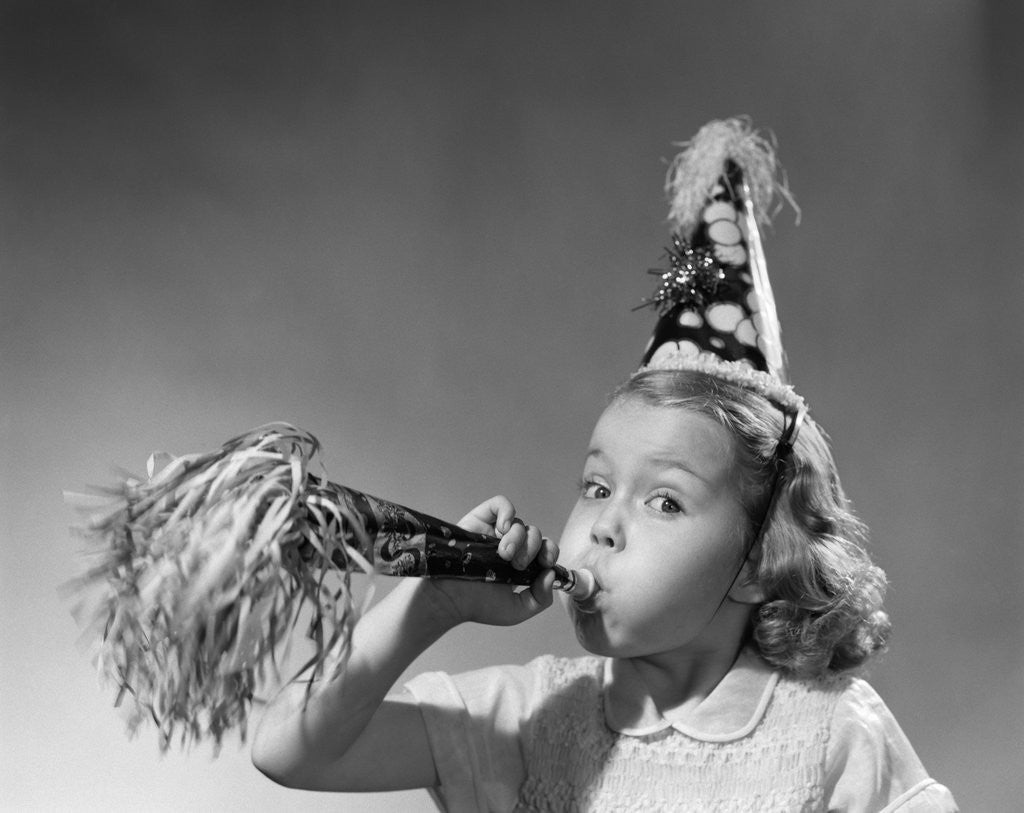 Detail of 1950s girl wearing party hat blowing into noise maker looking at camera by Corbis