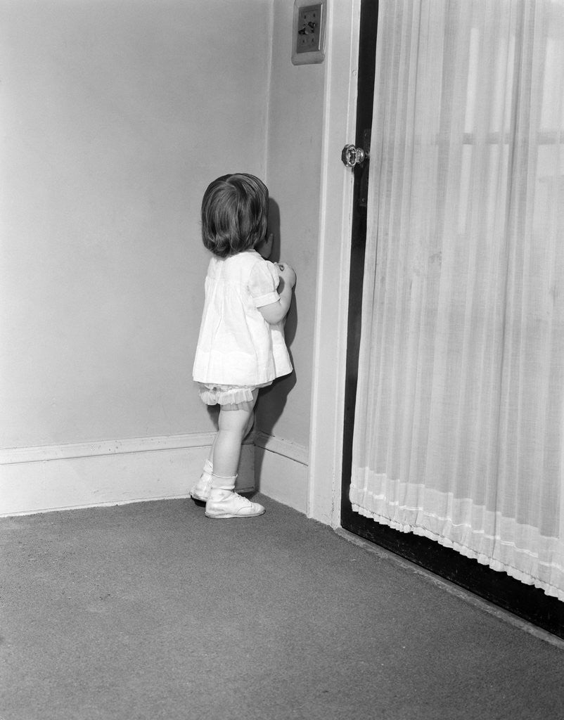 Detail of 1950s 1960s little girl in white dress being punished standing in a corner by Anonymous