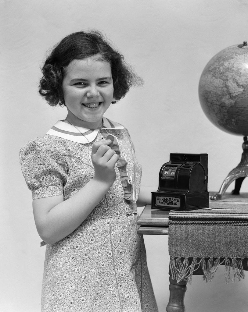 Detail of 1930s child smiling girl putting money coin into toy cash register bank by Corbis