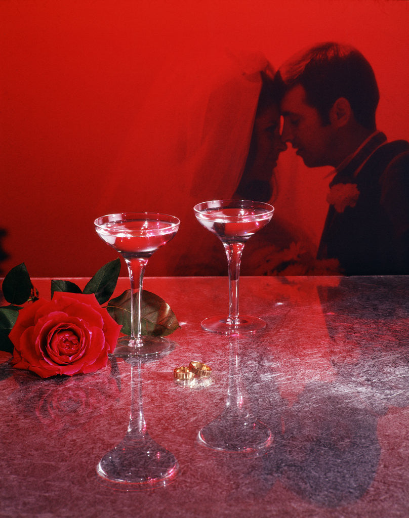 Detail of 1960s champagne flutes with rose and wedding rings in red filter with couple man woman bride groom in background by Anonymous