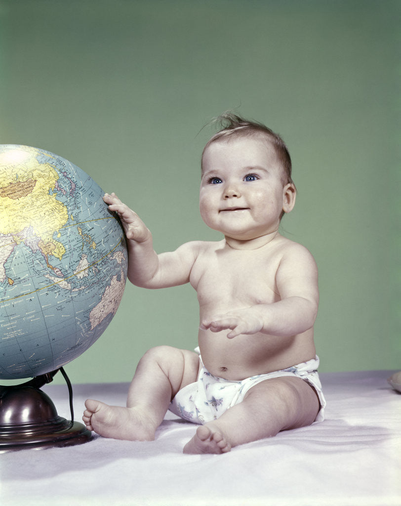 Detail of 1960s smiling baby girl sitting in diapers hand touching world globe by Anonymous