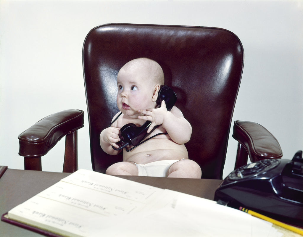 Detail of 1960s chubby baby sitting in leather chair at office desk holding telephone by Anonymous