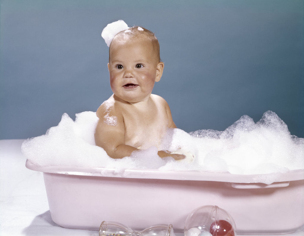 Detail of 1960s smiling baby sitting in bathtub bubble bath covered with soap bubbles by Anonymous