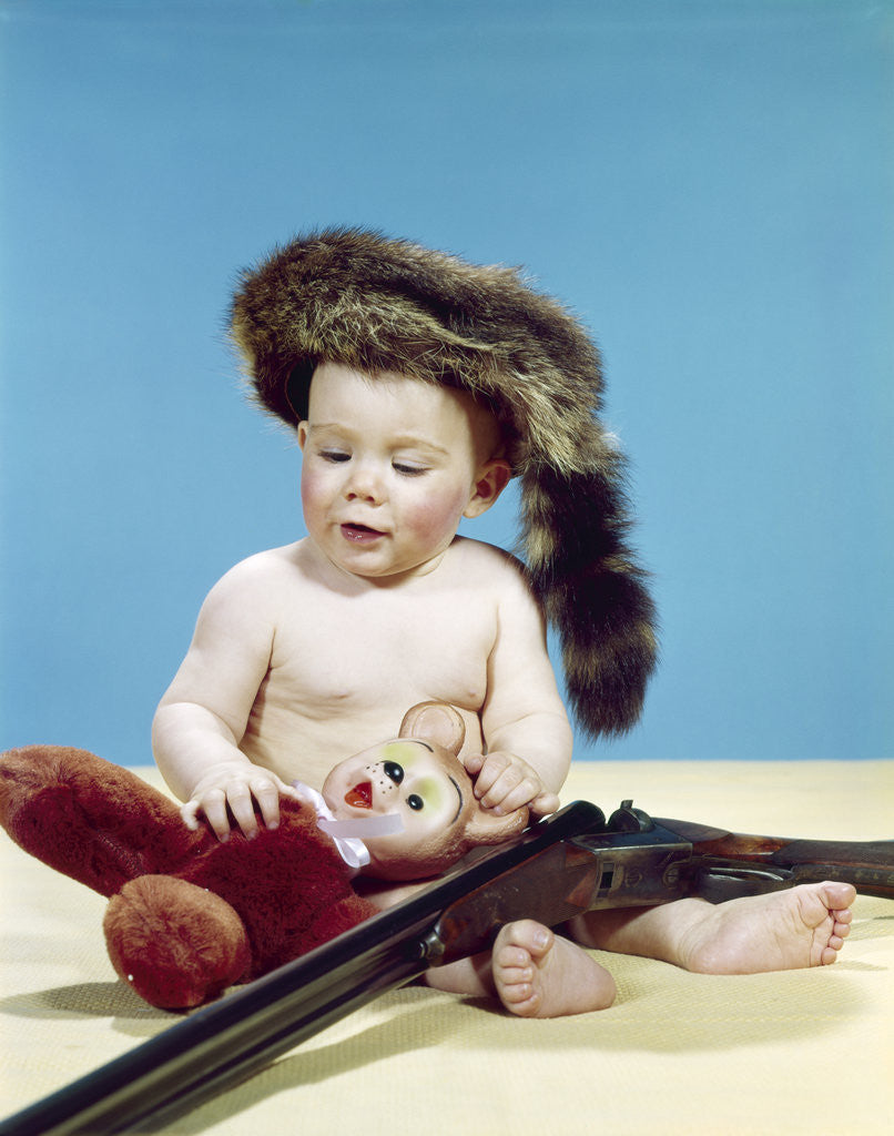 Detail of 1960s baby boy wearing coonskin cap with stuffed animal and shotgun by Anonymous
