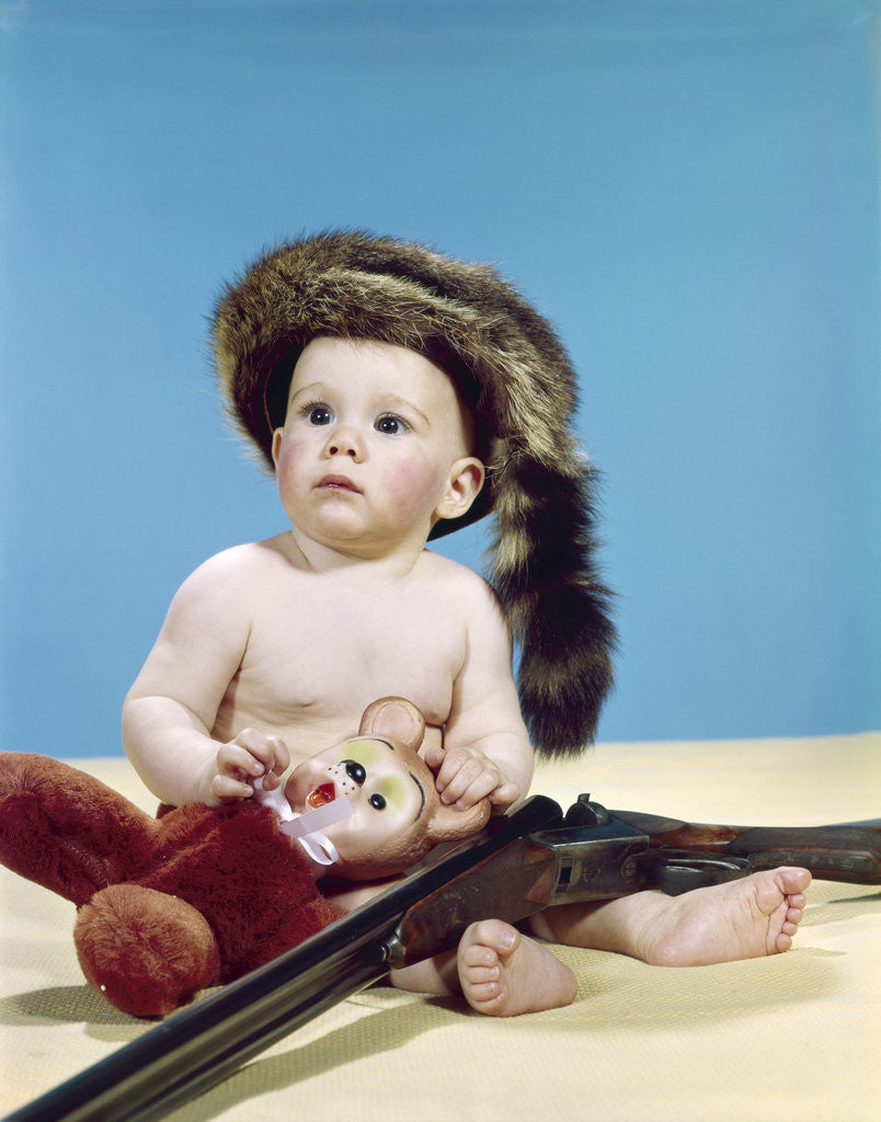 Detail of 1960s baby boy wearing coonskin cap with stuffed animal and shotgun gun by Corbis