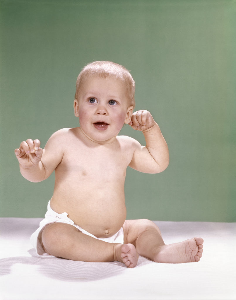 Detail of 1960s baby wearing diaper sitting on pink blanket mouth open hand touching ear by Anonymous