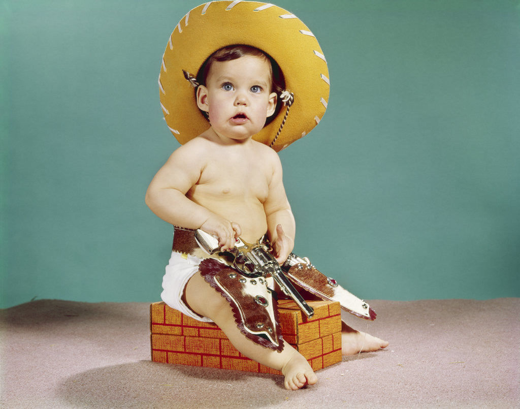 Detail of 1960s baby wearing cowboy hat and holster with guns pistols looking at camera by Anonymous