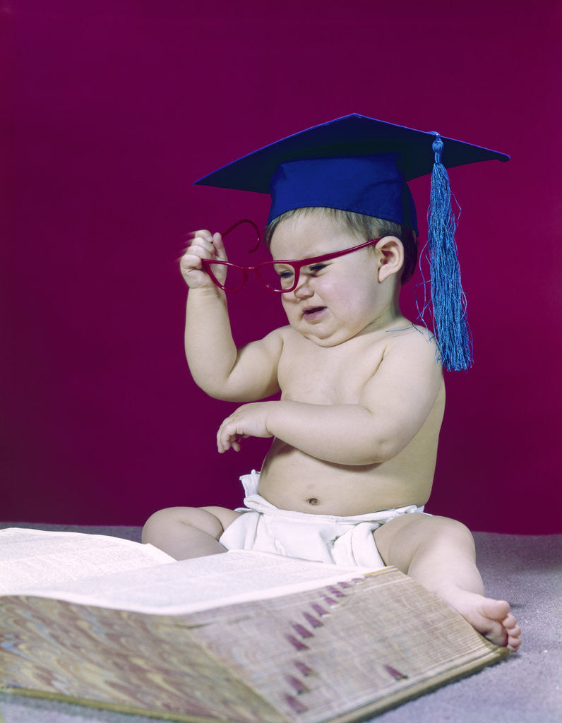 Detail of 1960s graduate baby wearing mortarboard eyeglasses reading book by Anonymous