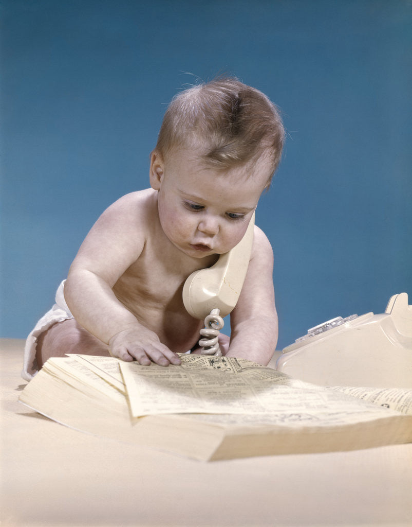 Detail of 1960s baby reading phone book yellow pages with telephone receiver on shoulder by Anonymous
