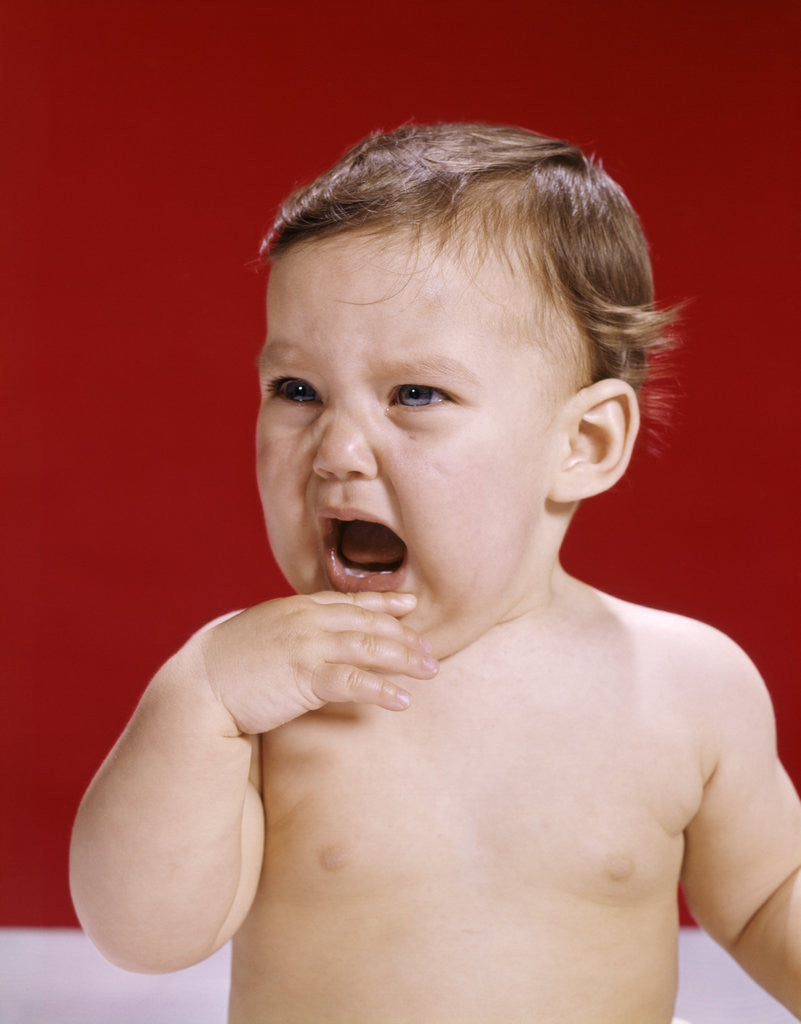 Detail of 1960s portrait of crying baby holding hand up to mouth red background by Anonymous