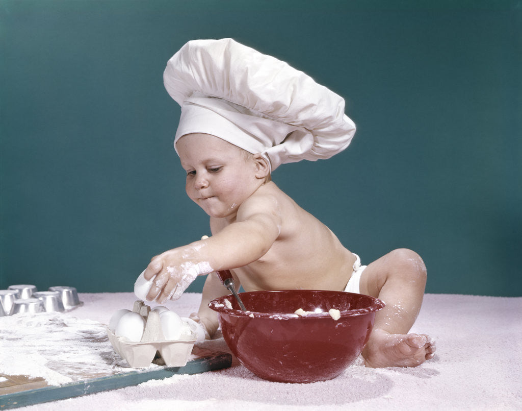 Detail of 1960s baby wearing chefs hat holding egg surrounded by cooking equipment by Anonymous