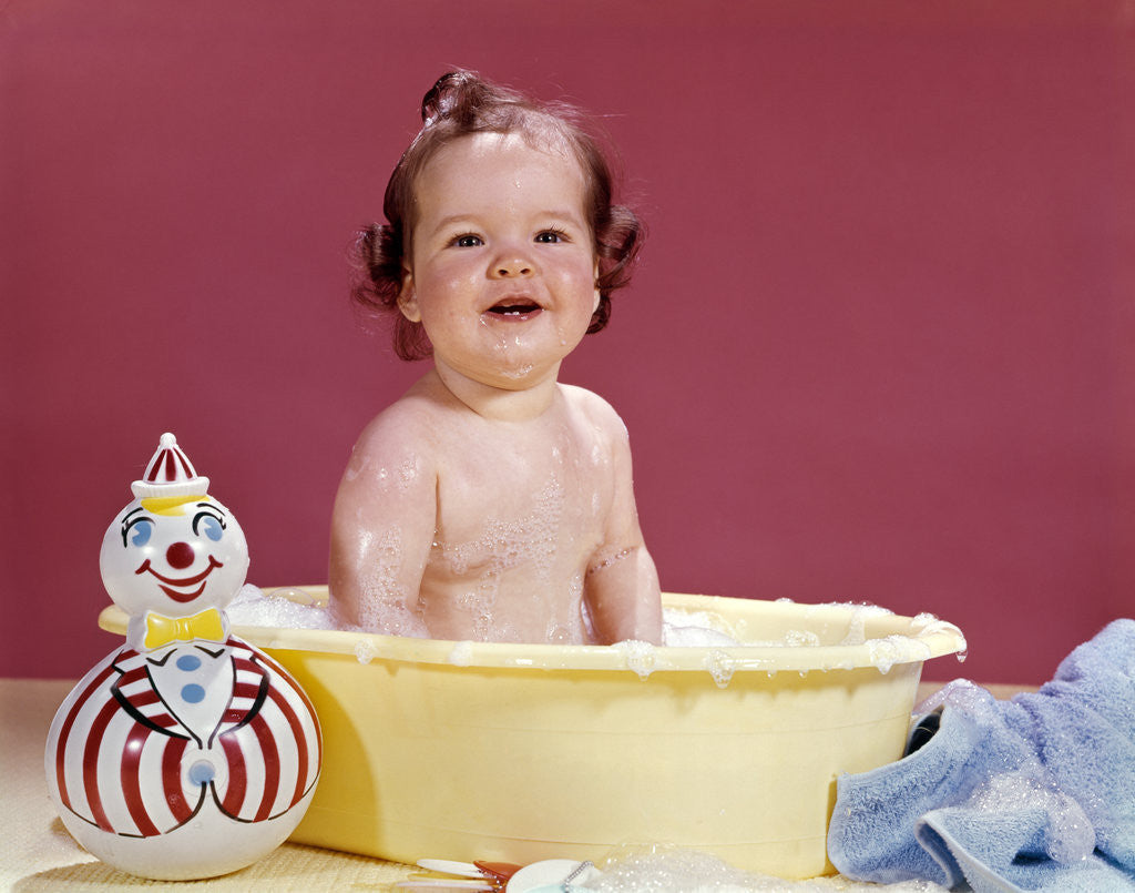 Detail of 1960s smiling brunette baby sitting in yellow plastic bath tub with toy clown looking at camera by Anonymous