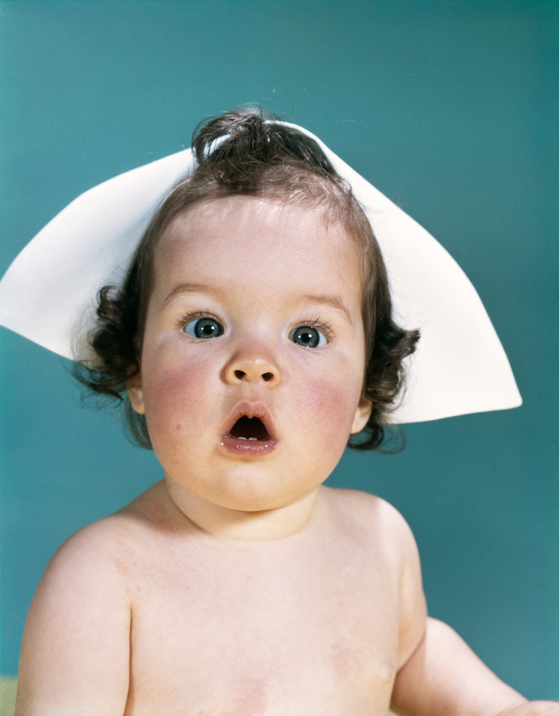 Detail of 1960s surprised baby facial expression wearing a nurse cap looking at camera by Anonymous