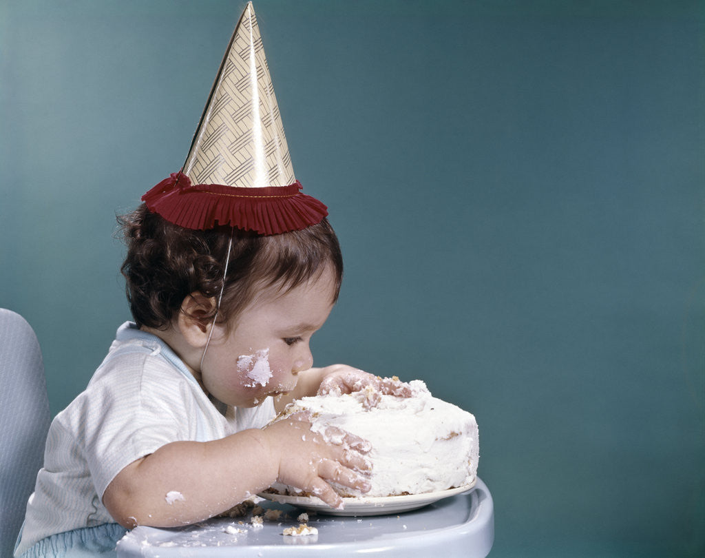Detail of 1960s baby in highchair wearing birthday hat eating whole birthday cake by Anonymous