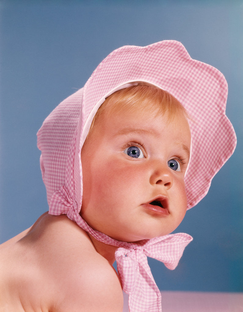 Detail of 1960s very cute blue eyed baby wearing pink white checked bonnet looking up by Anonymous