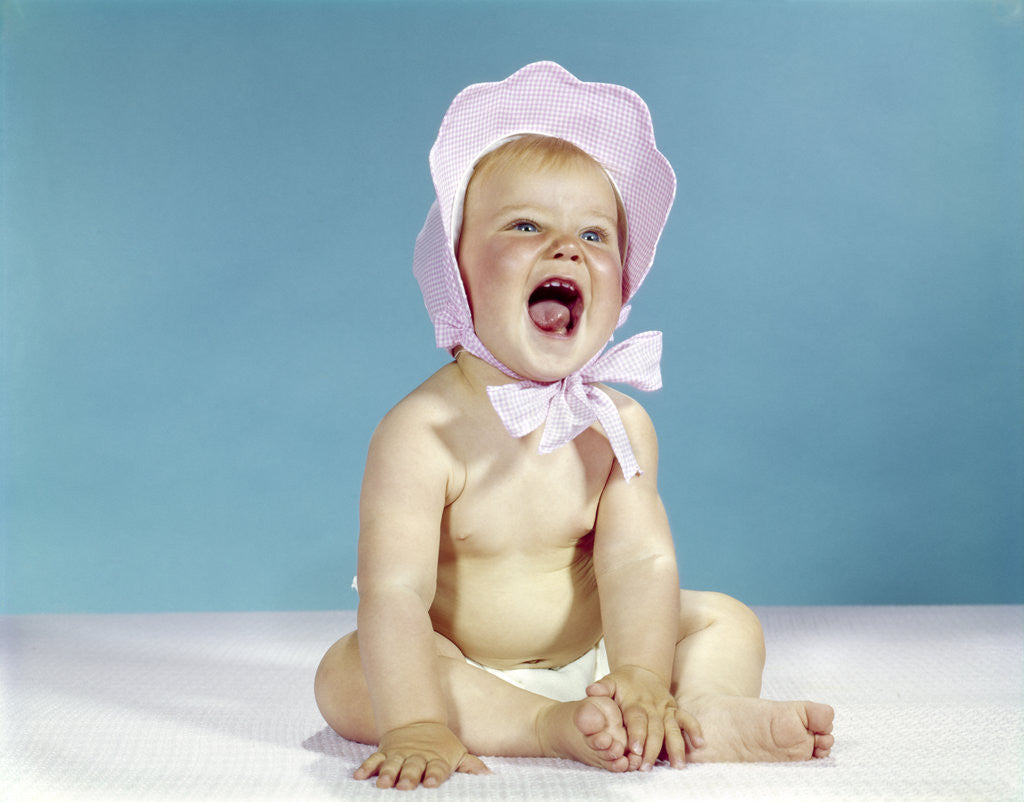 Detail of 1960s baby wearing pink checked bonnet laughing by Corbis