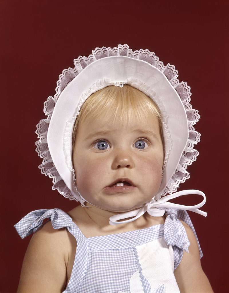 Detail of 1960s portrait baby girl wearing bonnet and blue gingham dress showing two front teeth looking at camera by Anonymous