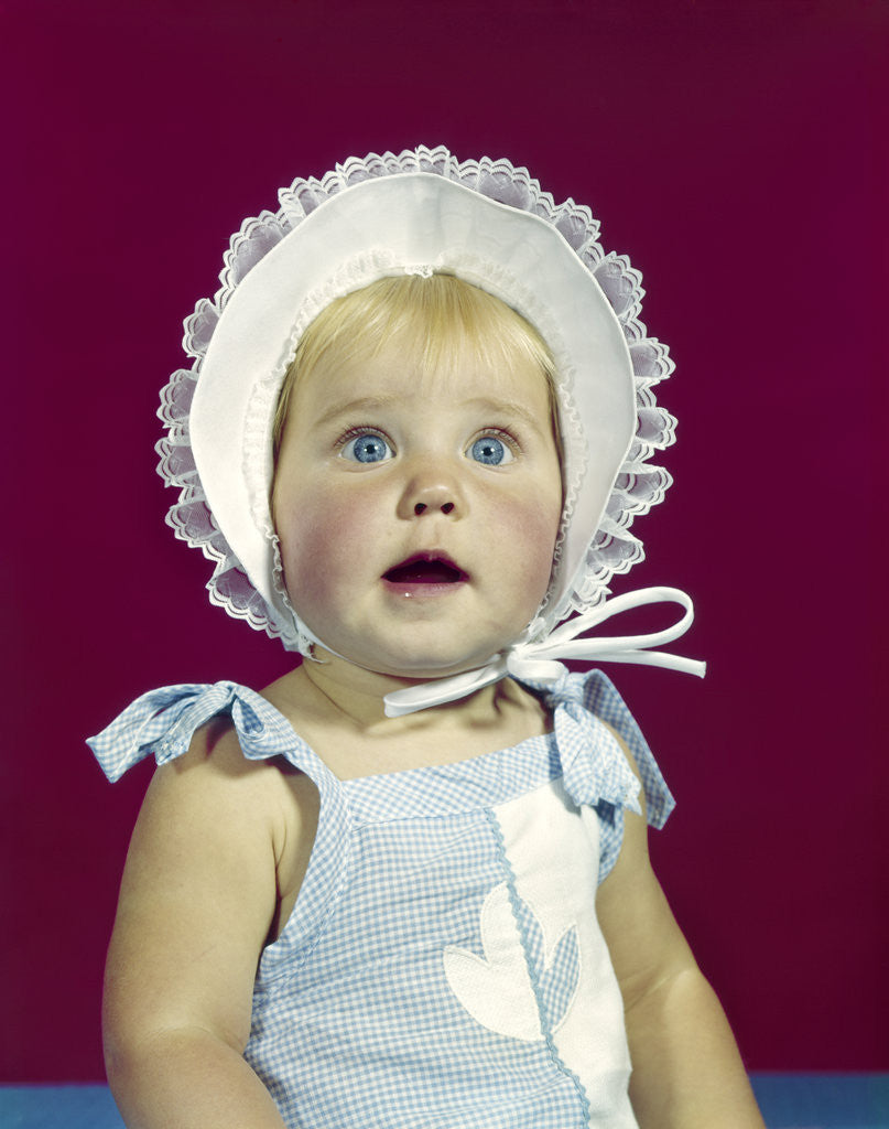 Detail of 1960s blonde baby blue eyes wearing ruffled bonnet facial expression of wonder looking at camera by Anonymous