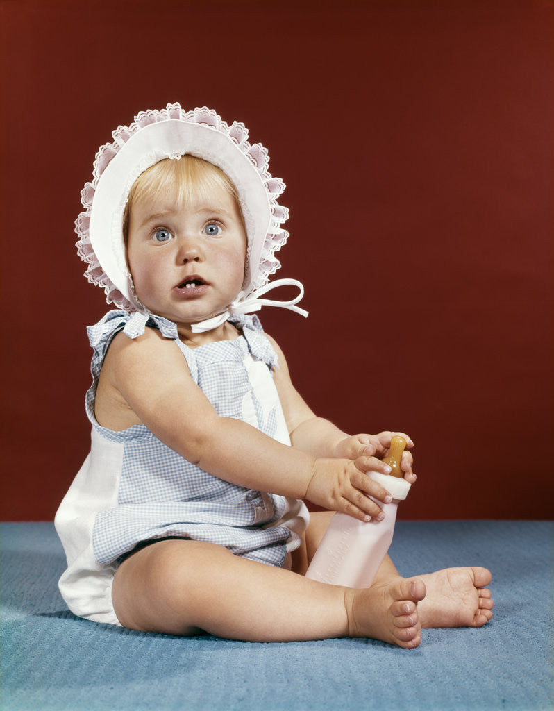 Detail of 1960s baby girl ruffled bonnet holding bottle funny facial expression looking at camera by Anonymous
