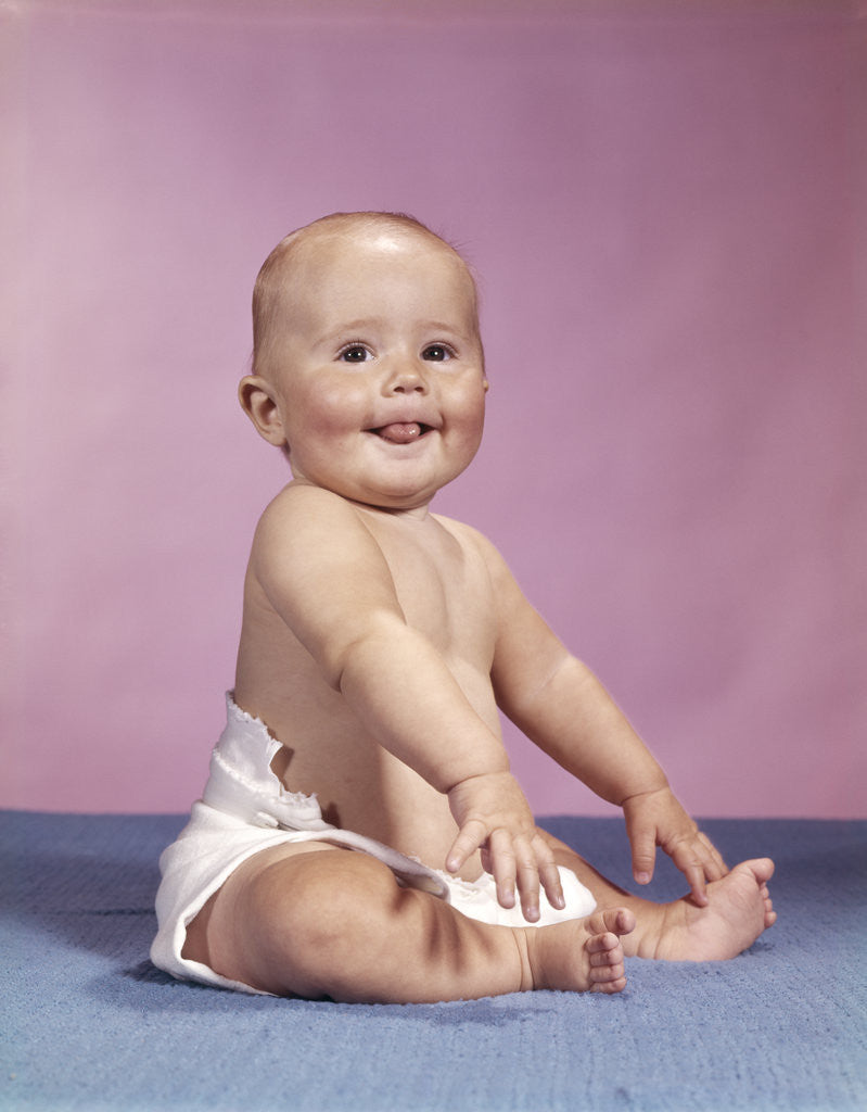 Detail of 1960s smiling baby wearing cloth diaper sitting on blue blanket sticking out tongue looking at camera by Corbis