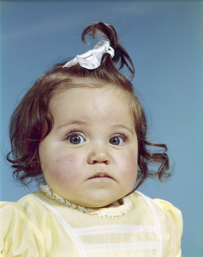 Detail of 1960s brunette baby girl ribbon topknot yellow shirt chubby cheeks round face sad unhappy facial expression looking at camera by Corbis
