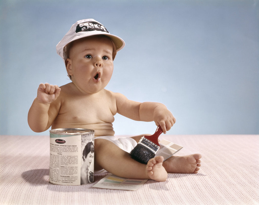 Detail of 1960s baby with funny facial expression wearing painters cap holding paintbrush sitting by paint can by Anonymous