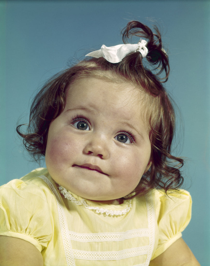 Detail of 1960s brunette baby girl round chubby face yellow dress ribbon topknot interested sincere facial expression looking at camera by Anonymous