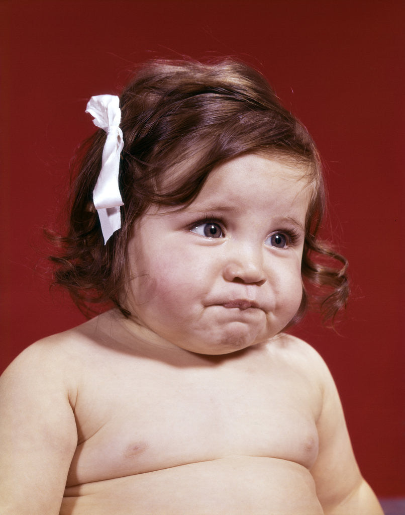 Detail of 1960s portrait of chubby unhappy baby girl with white ribbon bow in messy hair by Anonymous
