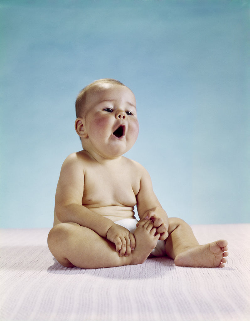 Detail of 1960s tired baby sitting holding on to one foot yawning facial expression by Anonymous