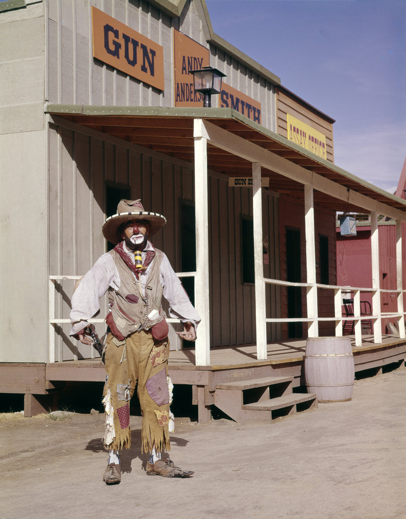 Detail of 1960s sad clown in cowboy costume standing in street of western frontier town by Anonymous