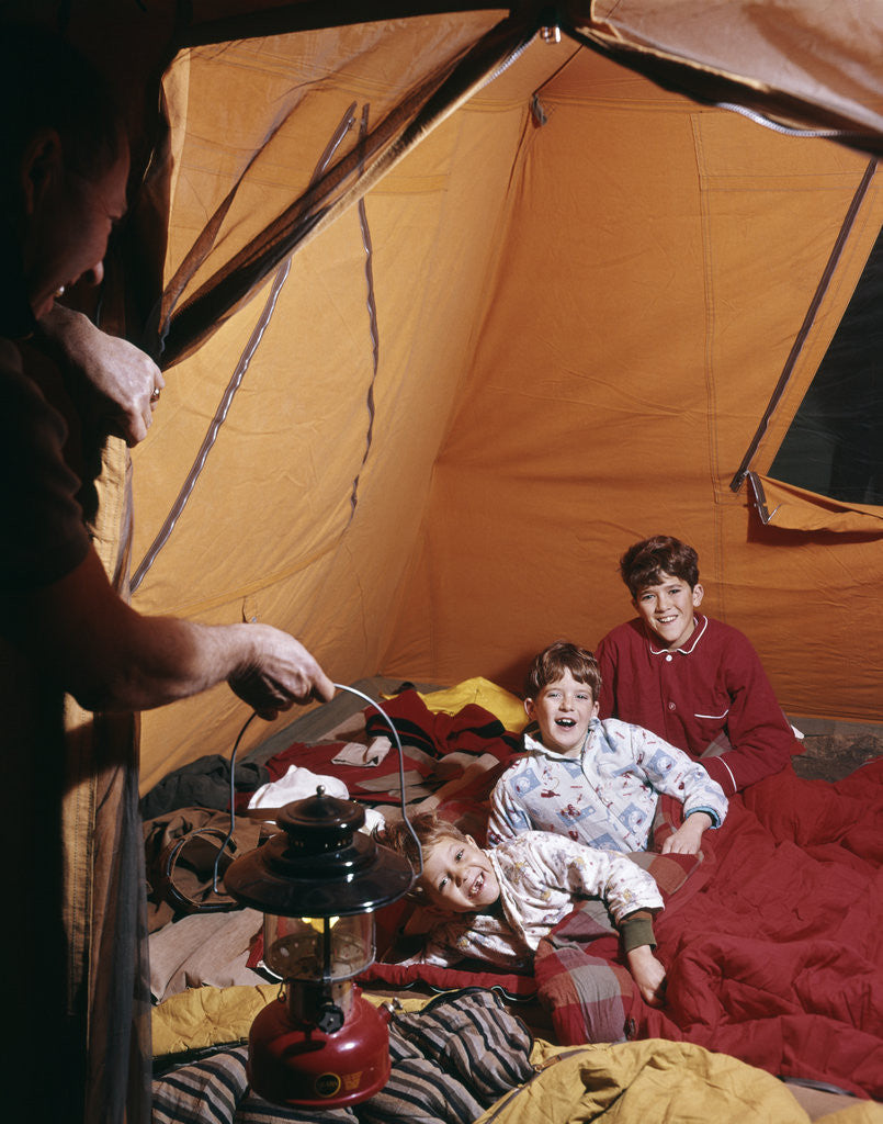 Detail of 1960s father with lantern checking up on 3 boys in sleeping bags pajamas in tent by Anonymous