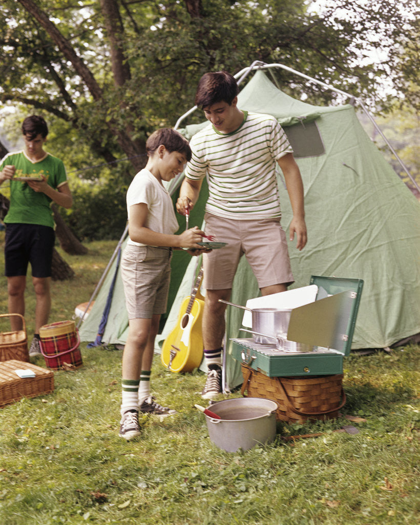Detail of 1960s three boys at campsite cooking camp stove eating food tent teens by Anonymous