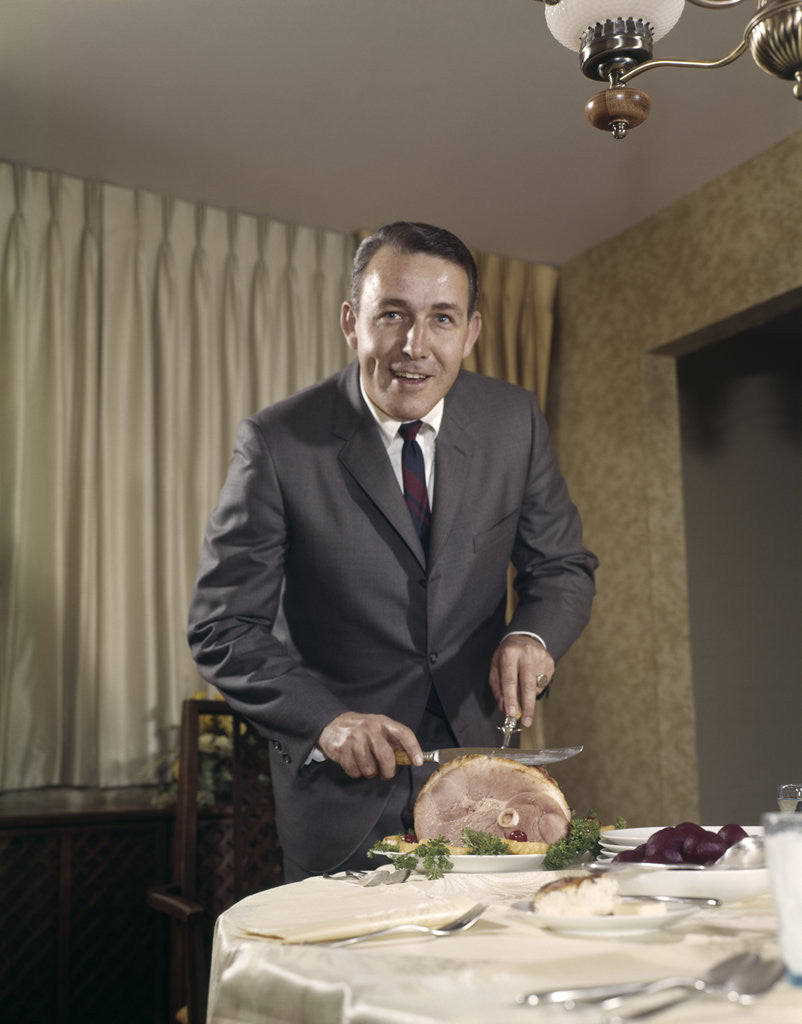 Detail of 1960s man carving ham dinner looking at camera by Corbis