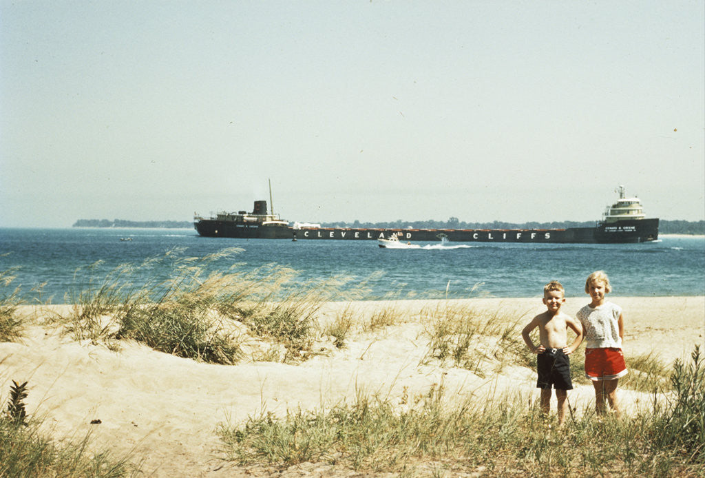 Detail of 1950s young boy and girl on beach smiling with a tanker ship on lake behind them port huron michigan 1955 by Anonymous