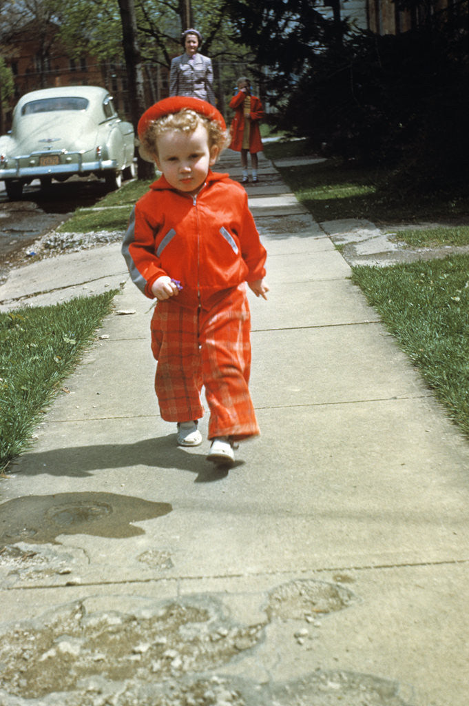 Detail of 1950s little boy in red outfit running on pavement with mother just behind by Corbis