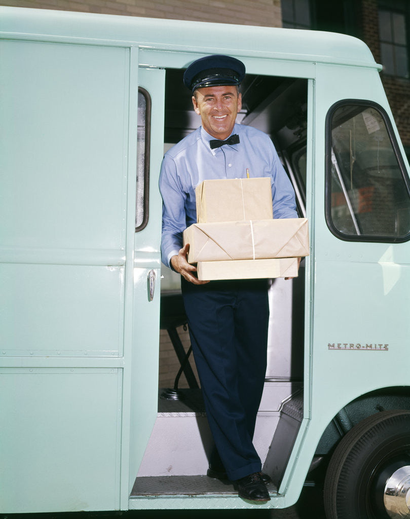Detail of 1950s 1960s man driver delivery truck van step out vehicle door holding packages boxes uniform hat service deliver work by Corbis