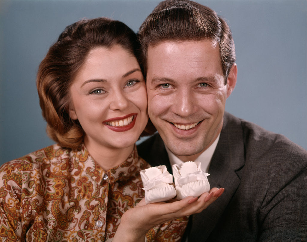 Detail of 1960s smiling couple holding baby shoes looking at camera by Anonymous