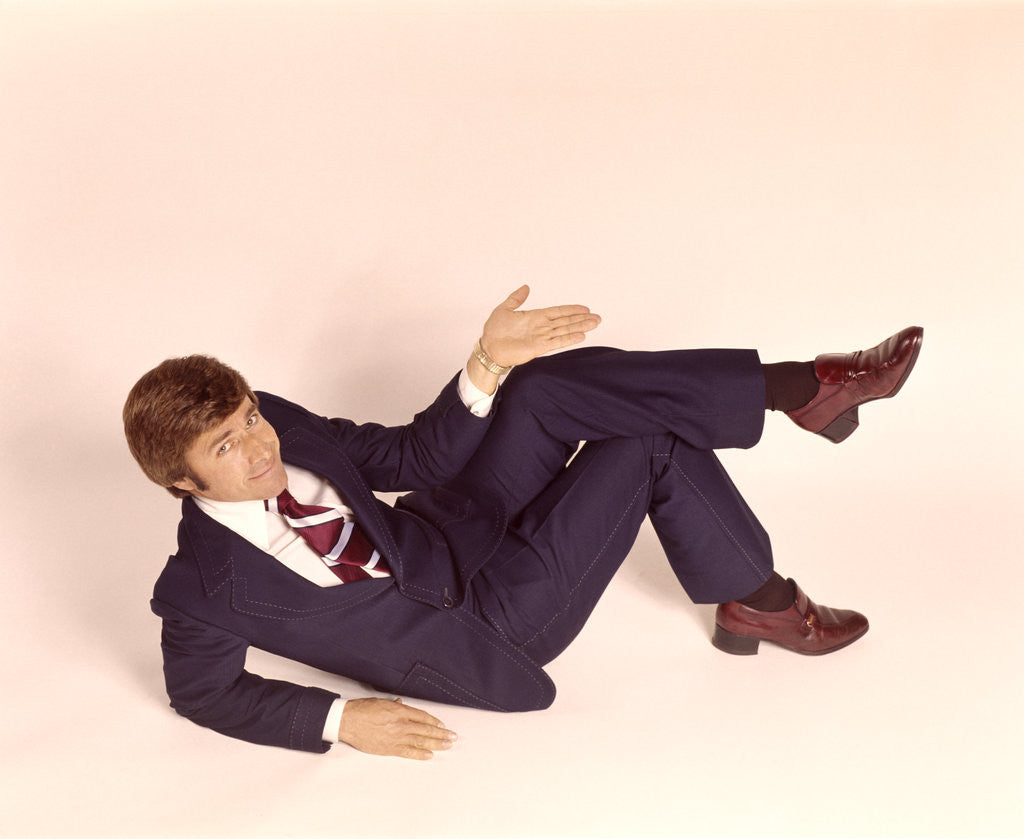 Detail of 1970s overhead looking down at man lying down legs crossed wearing business suit shrug off hand expression looking at camera by Anonymous