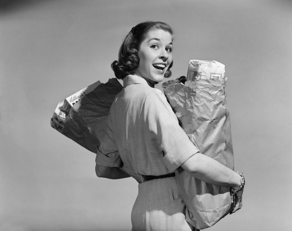 Detail of 1950s smiling woman carrying grocery bags both hands looking at camera over her shoulder by Anonymous